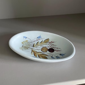 May include: Oval-shaped, off-white ceramic dish with a floral design. The design features brown, gold, and blue botanical elements. The dish is a decorative piece, suitable for serving or display.
