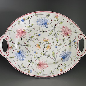 May include: Oval serving platter with handles. The white ceramic platter features a floral design with pink, blue, and yellow flowers, green leaves, and a red rim. The handles are white with a red trim.