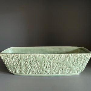 May include: A rectangular, light green ceramic planter with a textured, organic pattern. The planter has a slightly raised base and gently sloping sides. The overall design is simple yet elegant, suitable for various plants.