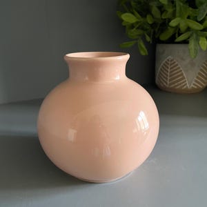 Könnte beinhalten: Eine kleine, kugelförmige Keramikvase mit einer glatten, glänzenden Oberfläche in einem sanften Pastellpfirsichton. Die Vase hat einen kurzen, schmalen Hals, der sich an der Öffnung leicht weitet und eine bauchige Form mit flachem Boden bildet.