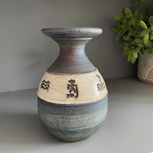 May include: A ceramic vase with a flared rim and a rounded body. The neck and base feature a dark, mottled blue and green glaze. A central band of cream-colored crackle glaze encircles the middle, decorated with three distinct dark brown symbols.