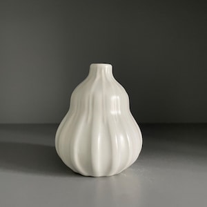 May include: A small, white ceramic vase with a bulbous, rounded body and a narrow neck. The surface of the vase features vertical, raised ridges that run from the base up to the neck, creating a textured, fluted appearance with a smooth finish.