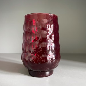May include: A ruby red glass tumbler with a textured, bubble-like pattern. The vintage-style drinking glass has a cylindrical shape and a solid base. The glass is approximately 5 inches tall.