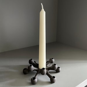 May include: A black metal candle holder with a distinctive design, holding a single white candle. The candle holder has a starburst shape with multiple circular openings for candles. The candle is tall and slender.