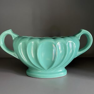 May include: A teal ceramic planter with two handles and a scalloped design. The planter is made of glazed ceramic and has a smooth, glossy finish.
