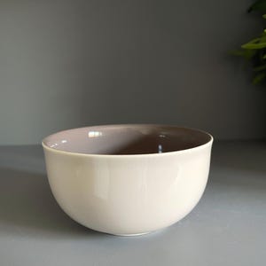 Könnte beinhalten: Eine runde Keramikschale mit einer glatten, glänzenden cremeweißen Außenseite und einem kontrastierenden matten taupefarbenen Innenraum. Die Schale hat eine einfache, geschwungene Silhouette mit einer weiten Öffnung und einem soliden Boden.