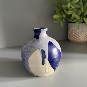 May include: A small, round ceramic vase with a narrow neck and a wide, bulbous body. The surface features a speckled light gray base with large, bold patches of deep blue glaze that drip down the sides, revealing areas of off-white clay.