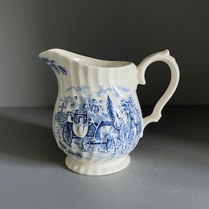 Puede incluir: Jarra de cerámica color crema con un diseño azul. La jarra presenta una escena detallada de un carruaje tirado por caballos y figuras. Tiene un asa curva y un pico decorativo, ideal para servir bebidas.