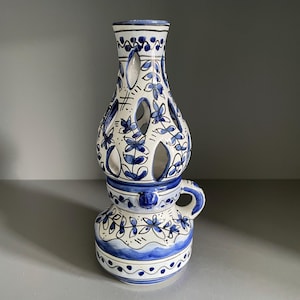 May include: A white and blue ceramic decorative lantern with a handle. The lantern has a cylindrical shape with cut-out sections and is decorated with blue floral patterns and a wavy border.