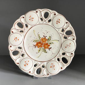 May include: White ceramic decorative plate with a scalloped edge and openwork design. The plate features a central floral motif in orange and green, with brown accents and small orange crosshatch details.