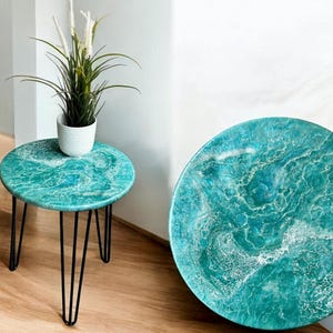 Könnte beinhalten: Zwei türkisfarbene und weiße Beistelltische aus Epoxidharz mit schwarzen Haarnadelbeinen. Auf einem Tisch steht eine kleine weiße Topfpflanze. Der andere ist eine passende Tischplatte. Die Tische haben ein marmoriertes, ozeanähnliches Design.