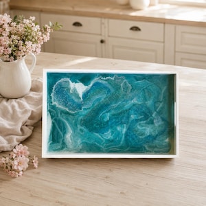 May include: A rectangular serving tray with a white frame and a glossy resin surface featuring swirling patterns of deep teal, turquoise, and white, mimicking the appearance of ocean waves with foam accents and a smooth, reflective finish.
