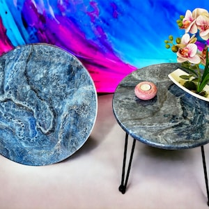 Puede incluir: Dos tapas de mesa redondas de mármol azul y gris con patas de metal negro. Una tapa de mesa tiene una vela rosa encima y la otra tiene una maceta de cerámica blanca con una planta verde y flores rosas.