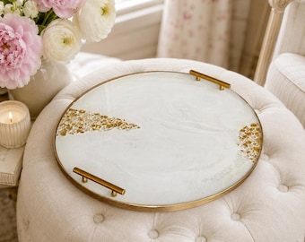 May include: A round serving tray with a white marbled resin surface, featuring embedded gold-toned metallic flakes and a thin gold-colored metal rim. The tray includes two horizontal gold-toned metal handles attached to opposite sides.