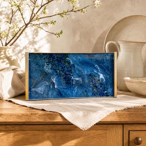 May include: A rectangular serving tray featuring a deep blue resin surface with swirling white patterns and embedded dark blue and gold-toned stone fragments. The tray is framed by thin metallic edges and has a smooth, glossy finish throughout.