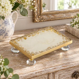 May include: A rectangular decorative tray featuring a smooth cream-colored center surface bordered by gold-toned metallic edges and embedded amber-colored glass fragments. The tray is supported by four clear, faceted crystal-style ball feet.