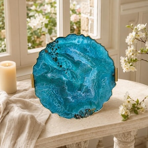 May include: A circular serving tray featuring a vibrant blue and teal resin surface with swirling patterns and a textured, geode-like appearance. The tray has a scalloped edge with a metallic gold finish and two matching gold handles on the sides.