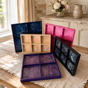 May include: A collection of five rectangular wooden display trays with multiple compartments. The trays feature colorful, marbled resin inserts in shades of deep blue, bright pink, light tan, dark purple, and black with white veining patterns.