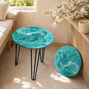 May include: A round side table with a teal and white swirling ocean-inspired resin top and three thin black metal hairpin legs. Beside the table stands a matching round resin disc featuring the same teal and white water-like pattern.