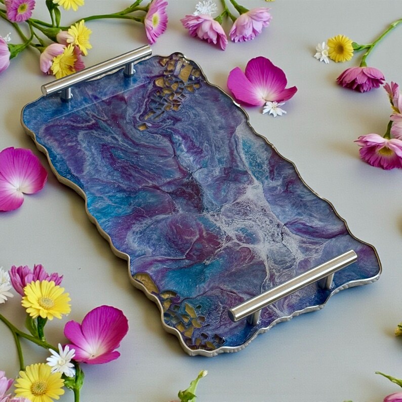 May include: A decorative serving tray with a unique, organic shape. The tray features a swirling pattern of blue, purple, and white resin, accented with gold details. It has silver handles and is surrounded by pink and yellow flowers.