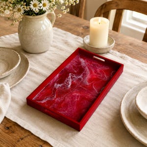 May include: A rectangular serving tray with a deep red and white marbled resin surface and a solid red frame. The tray features integrated cutout handles on the short ends and a glossy finish, showcasing swirling patterns of crimson and white.