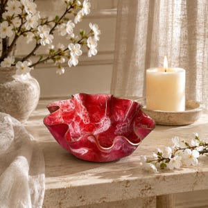 May include: A decorative bowl with a wavy, scalloped rim featuring a deep red and white marbled pattern. The bowl has a smooth, glossy finish and a gold-toned edge. It is positioned on a light-colored stone surface near a white pillar candle.