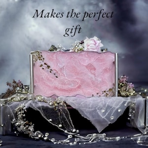 May include: A pink and white marbled box with a silver frame, decorated with a white sheer fabric and a pink flower. The box is sitting on a silver tray. The text "Makes the perfect gift" is written above the box.