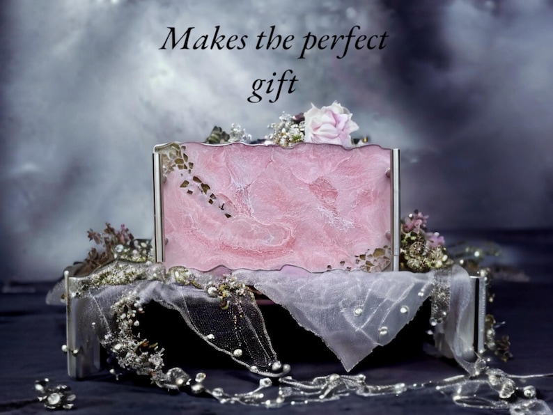 May include: A pink and white marbled box with a silver frame, decorated with a white sheer fabric and a pink flower. The box is sitting on a silver tray. The text "Makes the perfect gift" is written above the box.