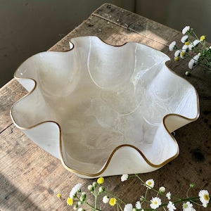 May include: A decorative white bowl with a wavy rim and a gold-colored trim. The bowl has a marbled pattern and is made of a translucent material. The bowl is empty and sits on a wooden surface. The bowl is a decorative item.