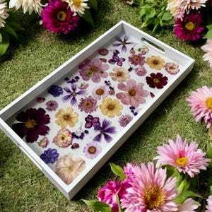 Puede incluir: Una bandeja de servicio rectangular blanca con asas, llena de una variedad de flores secas en tonos morados, rosas, amarillos y blancos. La bandeja está colocada sobre un lecho de hierba verde, rodeada de flores frescas.