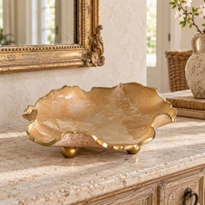 May include: A shallow, wide decorative bowl with a translucent, amber-toned, marbled surface and an irregular, wavy rim finished in gold. The bowl is supported by three small, spherical gold-colored feet, providing a stable base.