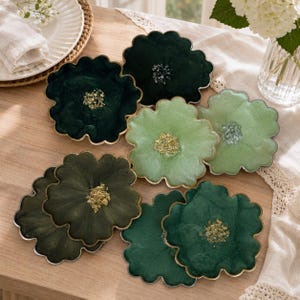 May include: A set of seven flower-shaped coasters in various shades of green, including dark forest green, olive, and light sage. Each coaster features a scalloped edge with a metallic trim and a textured, glittery center resembling a flower stamen.