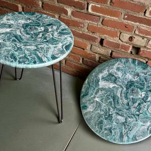 Könnte beinhalten: Zwei runde Beistelltische mit blaugrünen und weißen marmorierten Oberflächen. Ein Tisch hat schwarze Haarnadelbeine, der andere ist eine Tischplatte. Die Tische stehen vor einer Backsteinmauer.
