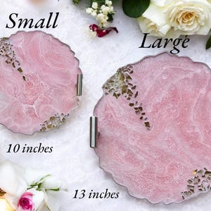 May include: Two pink resin serving trays with silver handles. The smaller tray is 10 inches in diameter and the larger tray is 13 inches in diameter. Both trays have a marbled design with gold flecks.