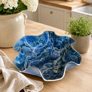 May include: A decorative bowl with a wavy, scalloped edge and a deep blue, marbled pattern featuring swirling white veins throughout the surface. The bowl has a smooth, glossy finish and a wide, open design, resting on a flat, wooden surface.