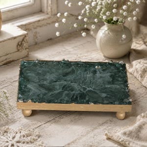 May include: A rectangular tray featuring a dark green, textured resin surface with a crystalline appearance. The tray is framed by a metallic gold edge and is supported by four small, rounded gold feet, providing a raised platform design.