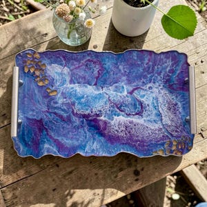 May include: A rectangular resin tray with a wavy edge, featuring a blend of blue, purple, and white colors. The tray has silver handles and is adorned with gold-colored accents. The tray is designed for serving or display.