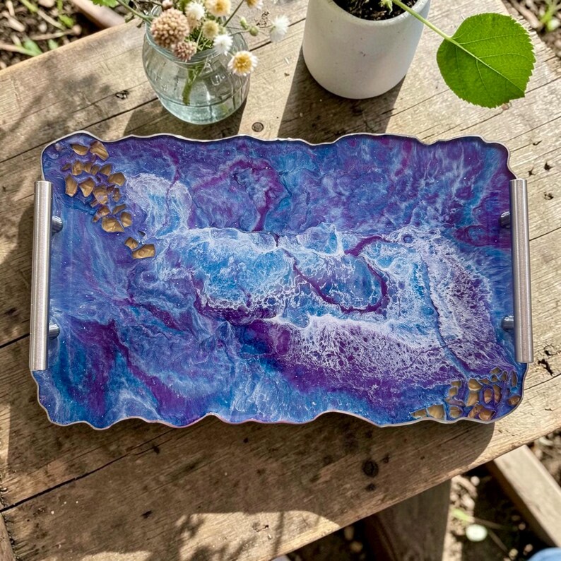 May include: A rectangular resin tray with a wavy edge, featuring a blend of blue, purple, and white colors. The tray has silver handles and is adorned with gold-colored accents. The tray is designed for serving or display.