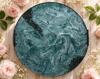May include: A round serving tray featuring a swirling teal and white marbled resin surface with black stone accents. The tray has a thin black metal rim and two black metal handles on opposite sides, creating a functional piece for serving.