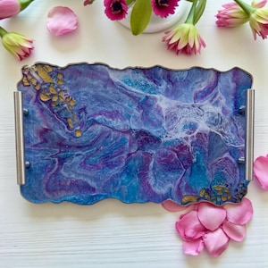 Puede incluir: Bandeja rectangular decorativa con un diseño único. La bandeja presenta un patrón arremolinado de resina azul, morada y blanca, acentuado con adornos dorados. Tiene asas plateadas y un borde ondulado.