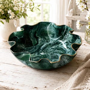 May include: A deep green bowl with a swirling marble-like pattern and a wavy, scalloped rim finished with a thin gold-toned edge. The bowl has a wide, shallow basin and a smooth, glossy surface that reflects light across its dark, textured interior.