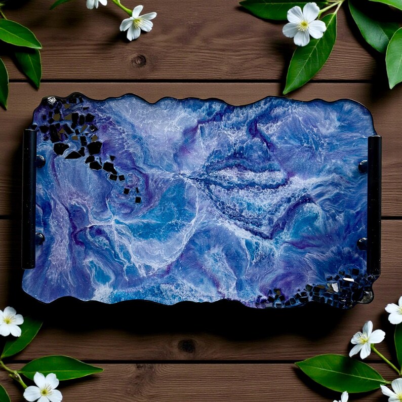 May include: A rectangular resin tray with a marbled blue and purple design. The tray has black handles and is decorated with black accents. White flowers and green leaves surround the tray on a wooden surface.