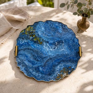 May include: A round, scalloped-edge serving tray featuring a swirling blue and white resin design with embedded gold-toned metallic fragments. The tray includes two gold-toned metal handles attached to opposite sides and a thin gold-toned rim.