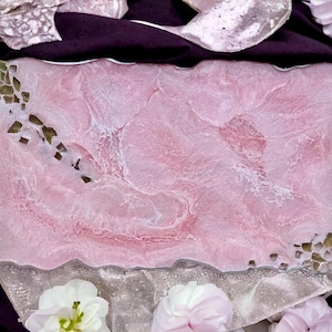 May include: A pink resin serving tray with a wavy edge and silver handles. The tray has a marbled design with white and light pink swirls.
