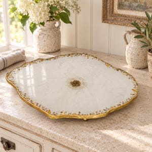 May include: A white, geode-inspired serving tray with a smooth, glossy surface, a textured center of small brown stones, and irregular, jagged edges finished with a metallic gold trim. The tray features a flat, circular design with gold accents.