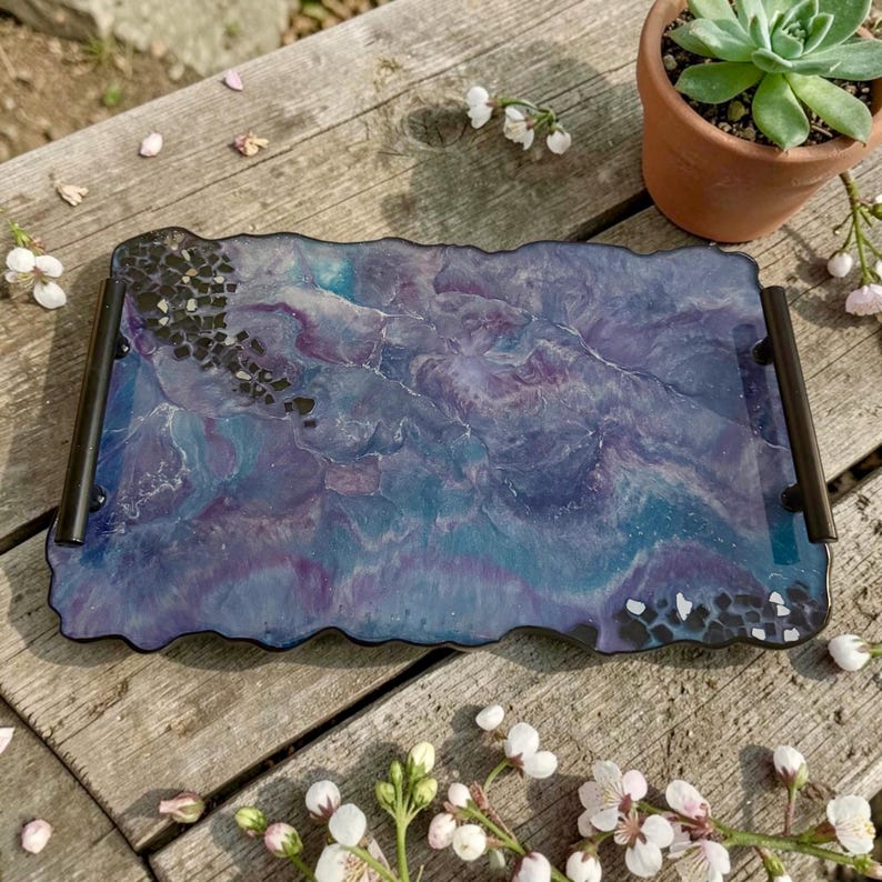 May include: A rectangular serving tray with a marbled design in shades of blue and purple. The tray has black handles and is decorated with black stones along the edges. A small succulent in a terracotta pot is in the background.