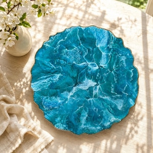 May include: A circular decorative tray featuring a vibrant blue and turquoise resin design that mimics the appearance of ocean waves with white foam accents. The tray has a scalloped, gold-toned metallic edge and a smooth, glossy finish.