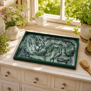 May include: A rectangular serving tray with a dark green frame and a marbled resin surface featuring swirling patterns of deep forest green, sage, and white, creating a textured, organic appearance reminiscent of a topographical landscape.