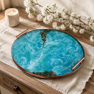 May include: A round serving tray featuring a swirling blue and turquoise resin surface with white foam-like patterns and dark stone accents. The tray is framed by a thin copper-colored metal rim and includes two matching copper-colored metal handles.