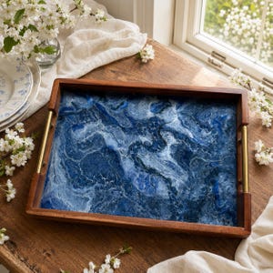 May include: A rectangular wooden serving tray featuring a deep blue and white marbled resin inlay. The tray has a smooth dark wood frame and two vertical gold-toned metal handles attached to the shorter sides, creating a functional serving piece.